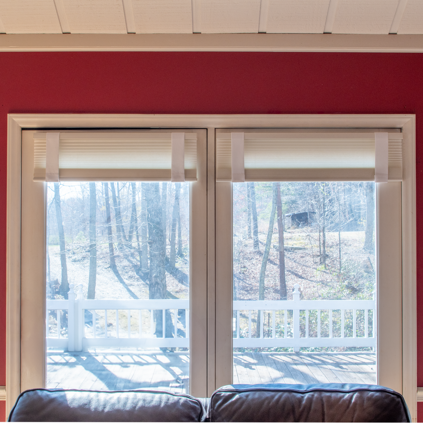 Living room windows with Lift Luxe Shade Straps in Cotton White securing pleated window shades. The crisp, structured straps keep the shades evenly aligned, framed by a bold red accent wall and soft natural light—perfect for enhancing blinds and shades with effortless elegance.