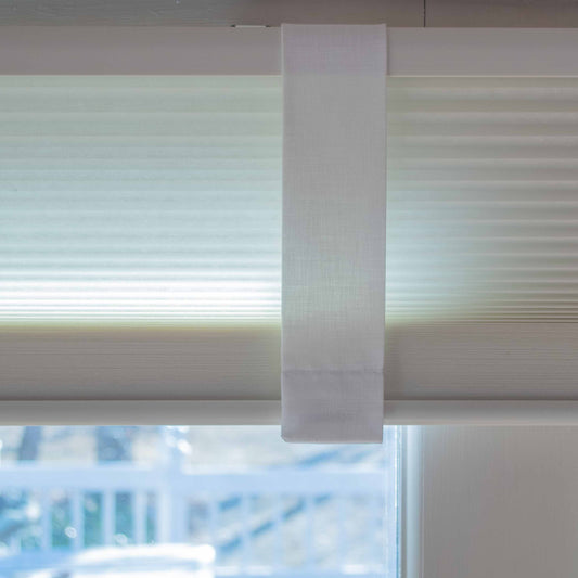 Close-up of a Lift Luxe Shade Strap in Cotton White securing a honeycomb window shade. The crisp, tailored strap blends seamlessly with the soft white fabric, ensuring a clean, structured look.
