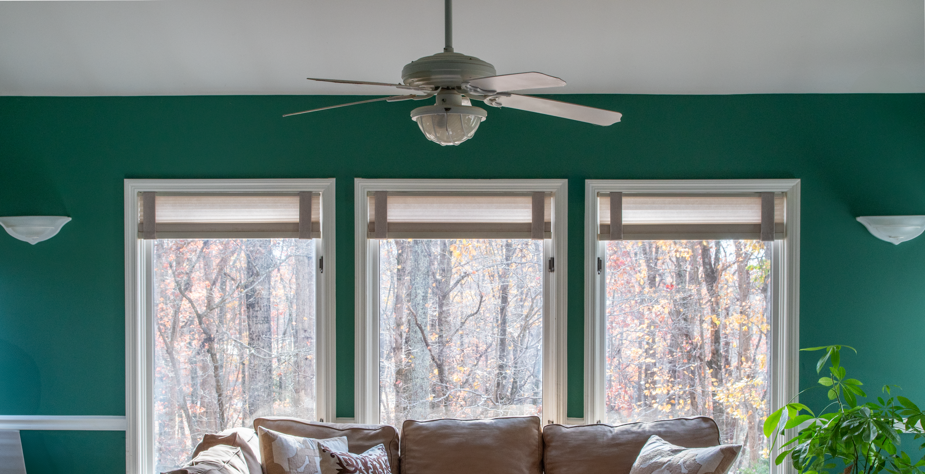 A stylish living room featuring three large windows framed by Lift Luxe Shade Straps in Linen Beige. The straps keep the cellular shades neatly aligned, complementing the earthy-toned sofa, textured pillows, and rich green walls for a cozy, polished space.