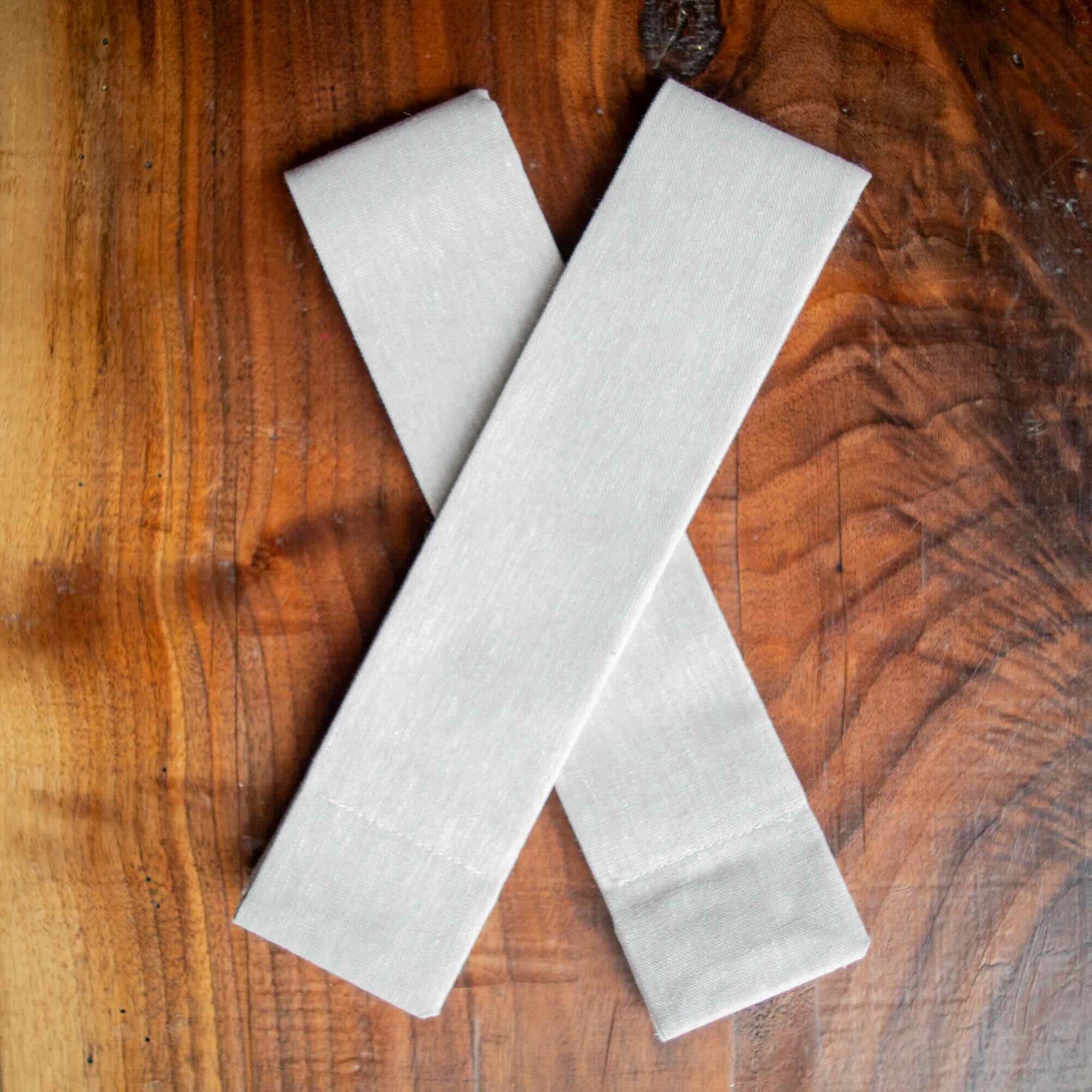 A pair of Lift Luxe Shade Straps in natural linen color crossed neatly on a polished wooden surface, highlighting the fabric’s texture and craftsmanship.