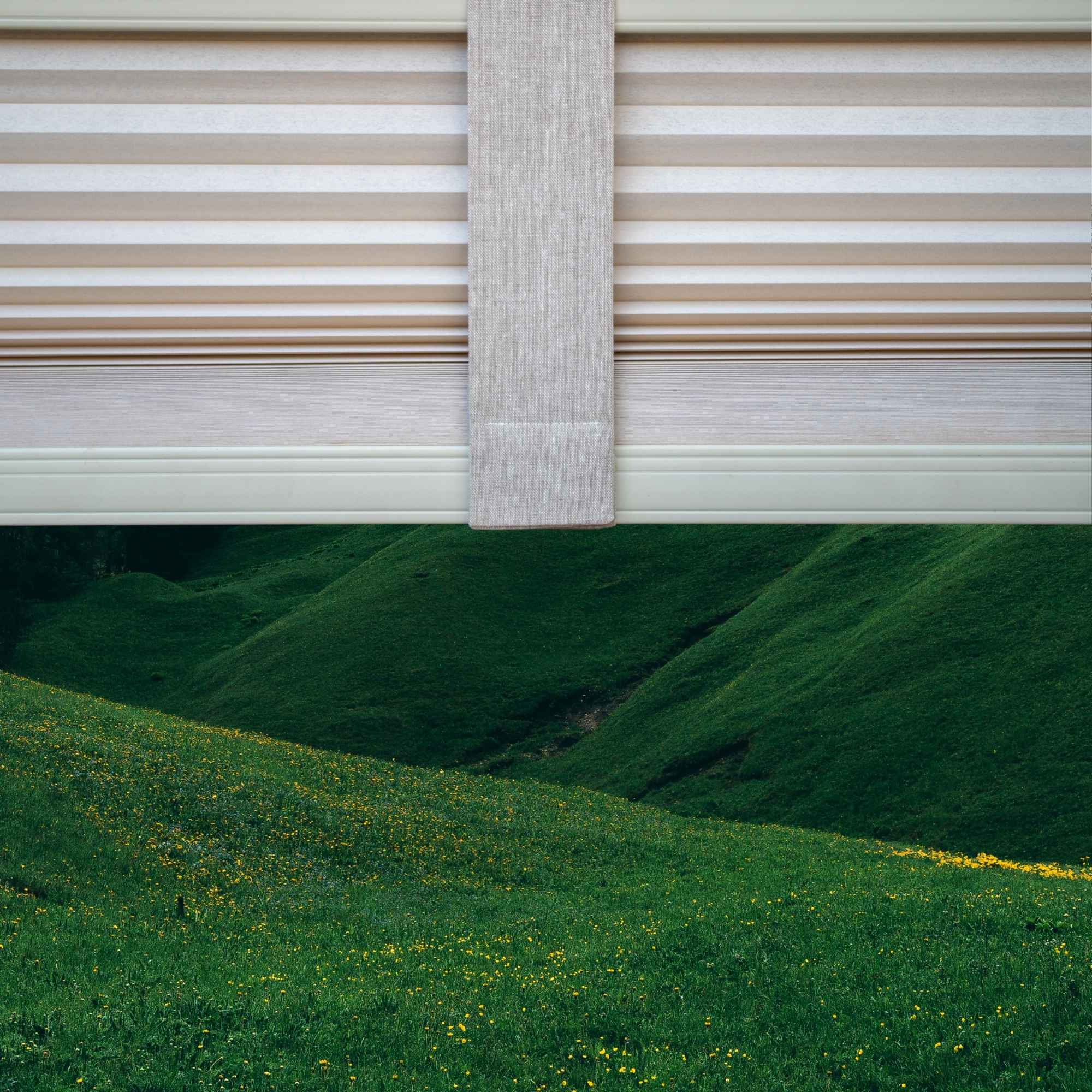 A Linen Beige Lift Luxe Shade Strap straightening a pleated window shade, framing a serene landscape of rolling green hills dotted with yellow wildflowers. The soft, natural fabric complements the organic scenery, creating a seamless balance between structured design and nature’s beauty.
