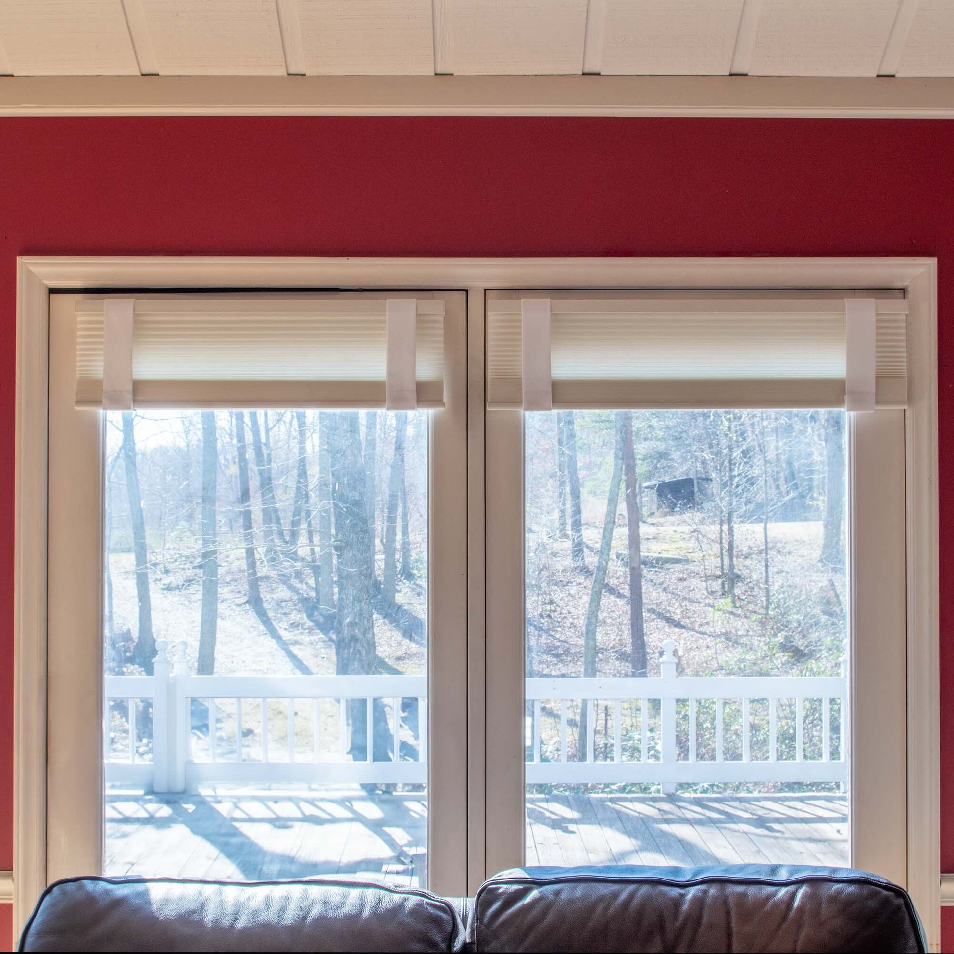 Two large living room windows with white Lift Luxe Shade Straps securing pleated blinds, framed by a red accent wall and overlooking a wooded backyard with deck railing.