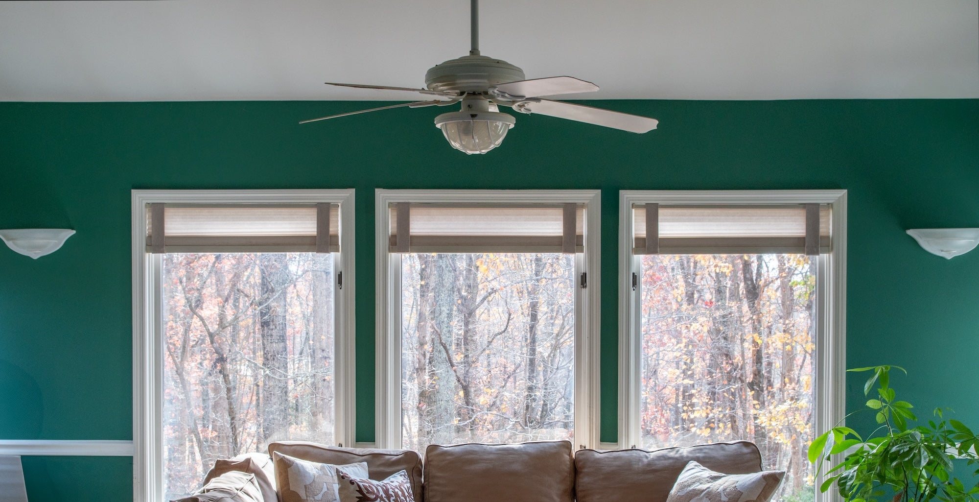 A stylish living room featuring three large windows framed by Lift Luxe Shade Straps in Linen Beige. The straps keep the cellular shades neatly aligned, complementing the earthy-toned sofa, textured pillows, and rich green walls for a cozy, polished space.