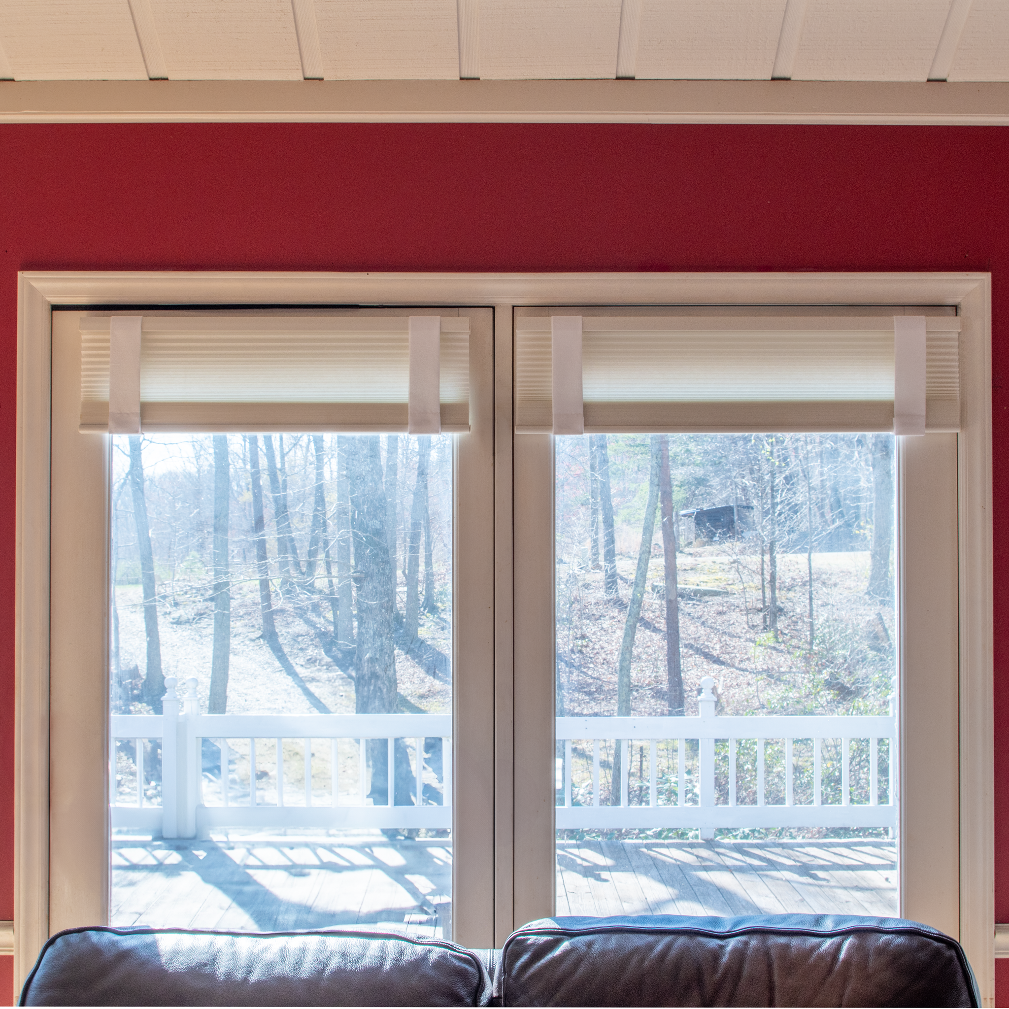 Living room windows with Lift Luxe Shade Straps in Cotton White securing pleated window shades. The crisp, structured straps keep the shades evenly aligned, framed by a bold red accent wall and soft natural light—perfect for enhancing blinds and shades with effortless elegance.